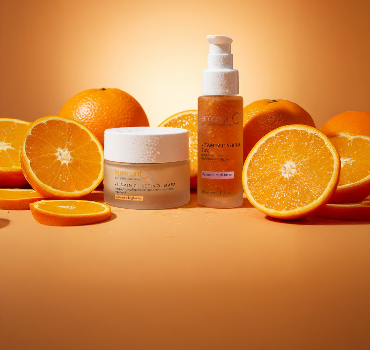 Two professional luxury skin care products are displayed among whole and sliced oranges on an orange background. The EmerginC Vitamin C Retinol Mask and Vitamin C Serum in a pump bottle emphasize a vibrant citrus theme.