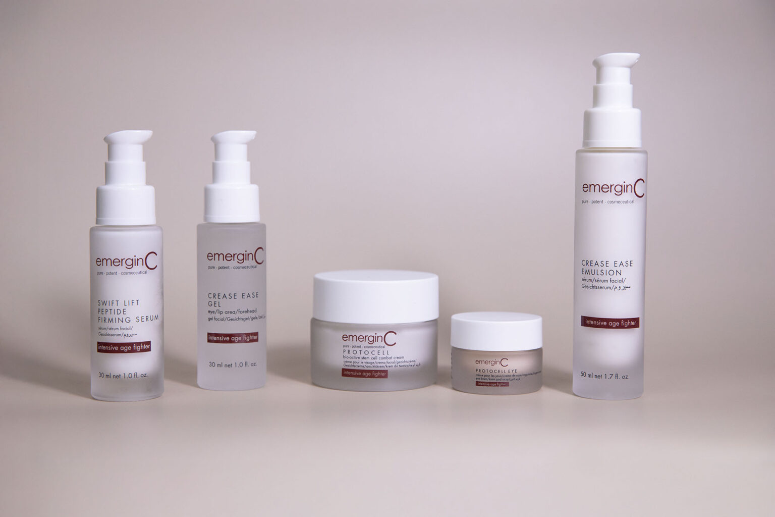 Five emerginC skincare products from the "Precise Age Fighter" line are arranged in a row against a plain, light beige background. Packaging includes pump bottles and jars with white and red labels.