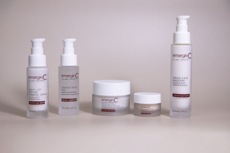 Five emerginC skincare products from the "Precise Age Fighter" line are arranged in a row against a plain, light beige background. Packaging includes pump bottles and jars with white and red labels.