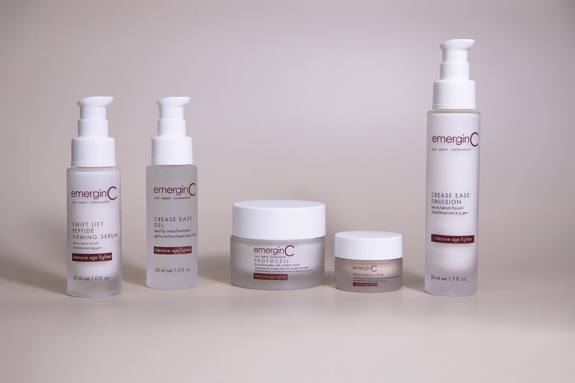 Five emerginC skincare products from the "Precise Age Fighter" line are arranged in a row against a plain, light beige background. Packaging includes pump bottles and jars with white and red labels.