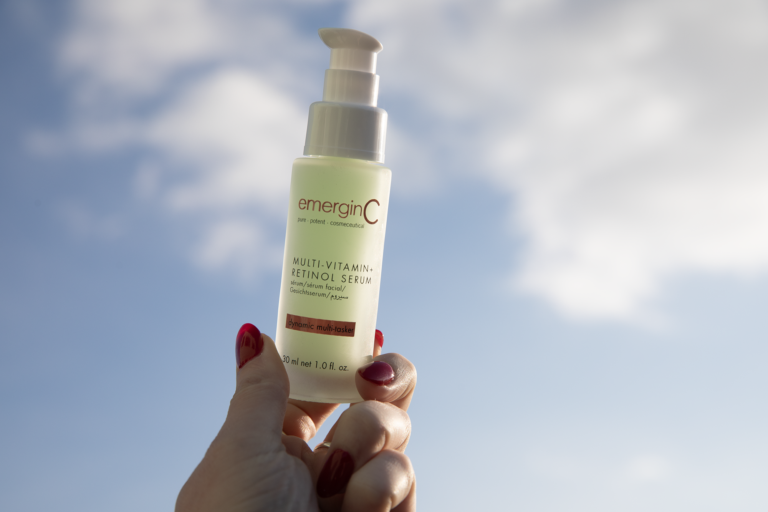 A hand with red nail polish holds a bottle of emerginC Multi-Vitamin+ Retinol Serum against a cloudy sky.