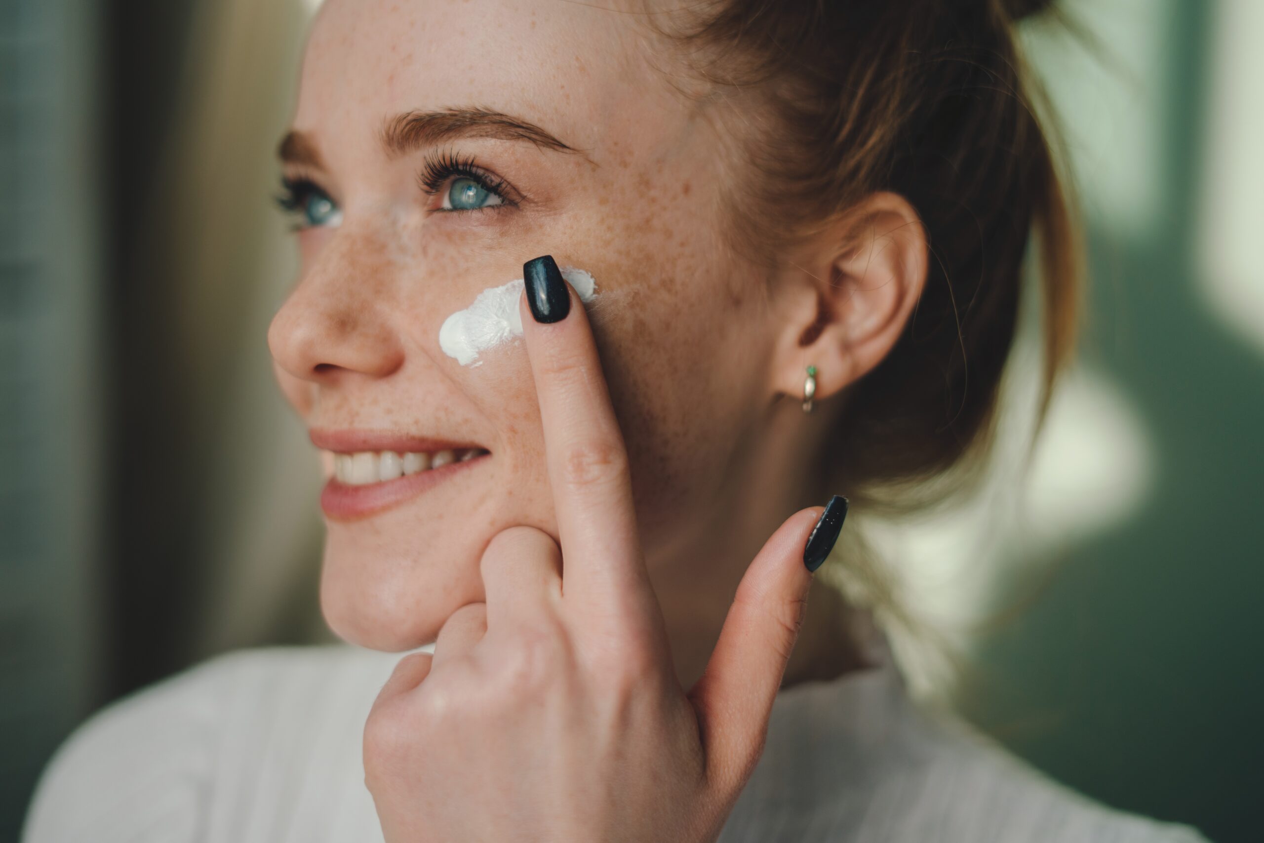 A smiling person with freckles applies a dab of cream to their cheek with their finger. They have blue eyes, light skin, and are wearing a white top. Their nails are painted dark, and they have a small hoop earring.
