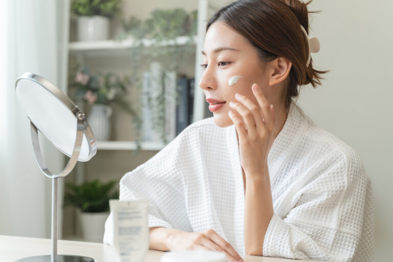 A woman in a white bathrobe applies face cream while looking into a round mirror. Surrounded by natural skin care for sensitive skin, she sits at a table in a bright, cozy room filled with plants and shelves.