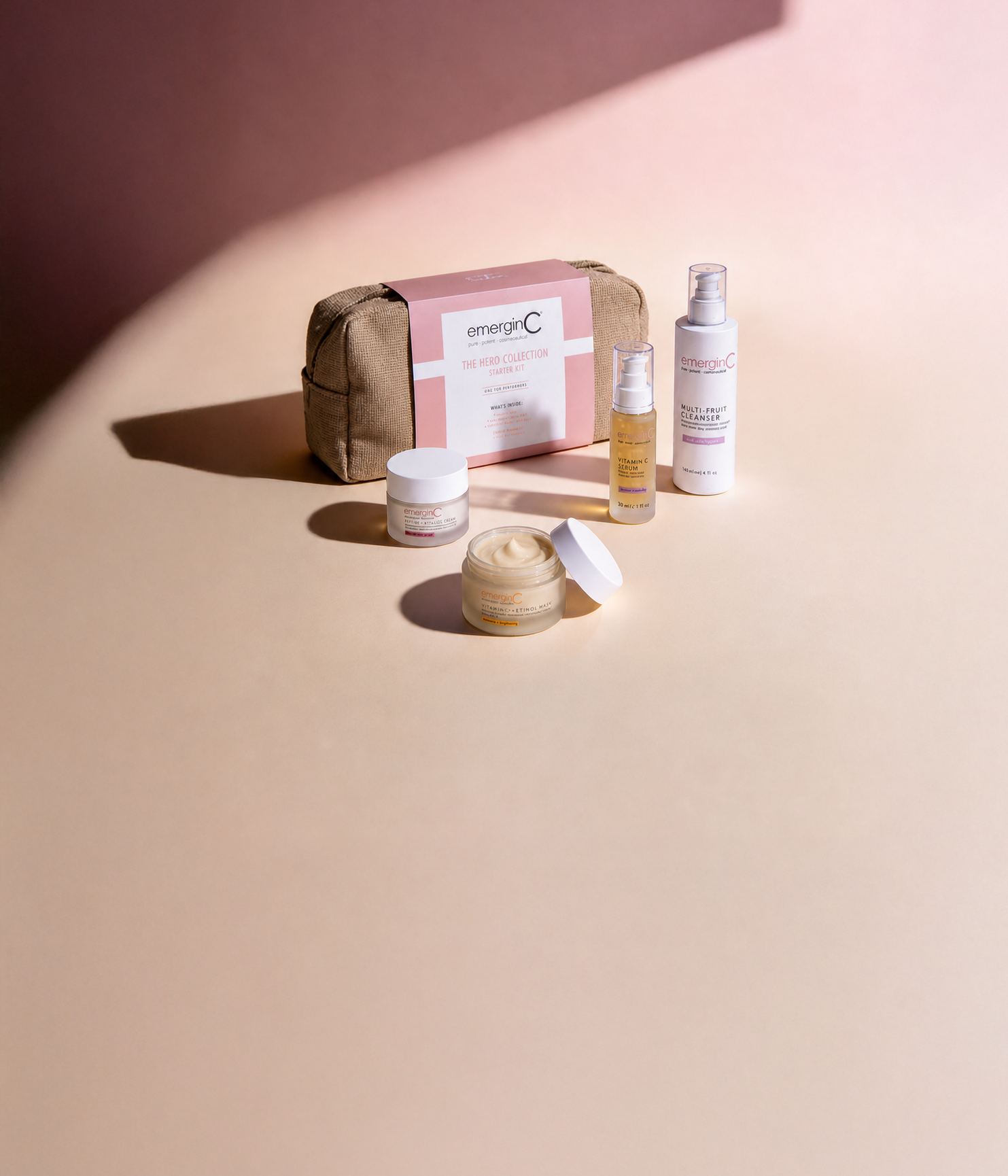 A collection of professional luxury skin care products from EmerginC, including a wrapped kit, two small jars, and two bottles, all arranged neatly on a smooth light pink and beige surface with a shadow in the background.
