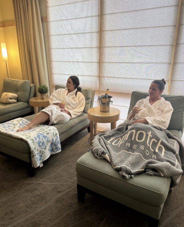 Two women in white robes relax on lounge chairs in a cozy spa setting. One has a blanket reading "Topnotch Resort," both hold drinks, with a champagne bottle in an ice bucket nearby. Soft lighting and curtains create a calm atmosphere.