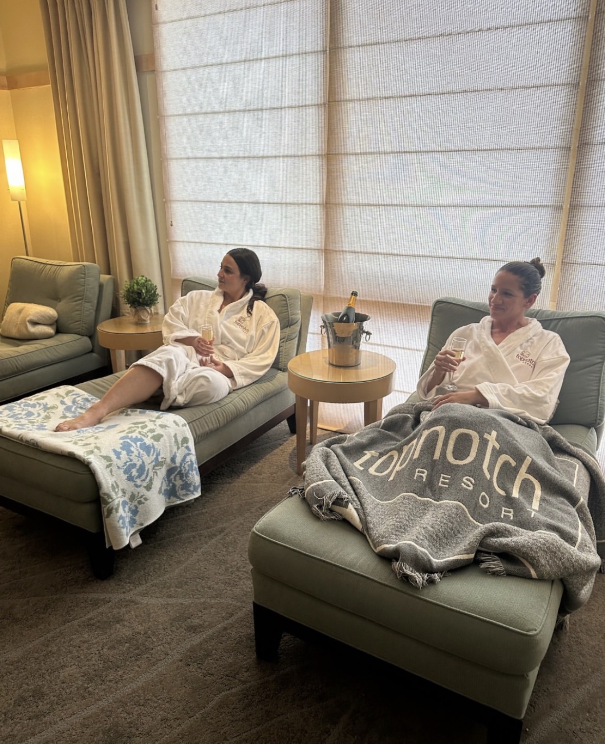 Two women in white robes relax on lounge chairs in a cozy spa setting. One has a blanket reading "Topnotch Resort," both hold drinks, with a champagne bottle in an ice bucket nearby. Soft lighting and curtains create a calm atmosphere.