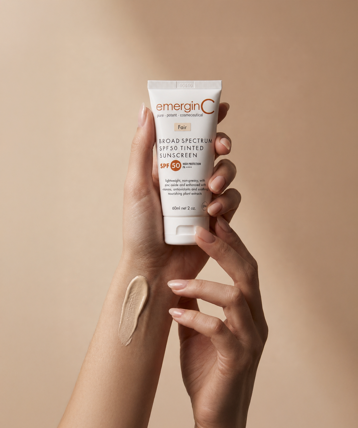 A hand holds a tube of emerginC SPF 50 tinted sunscreen, a professional luxury skin care essential. Another hand displays a swatch of the product on the forearm against a neutral beige background.