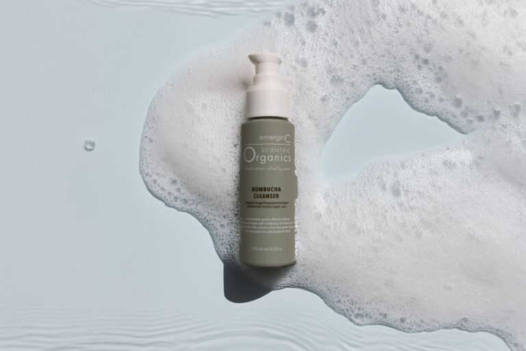 A bottle of EmerginC Scientific Organics Kombucha Cleanser is placed upright on a light blue surface, surrounded by white, foamy bubbles.
