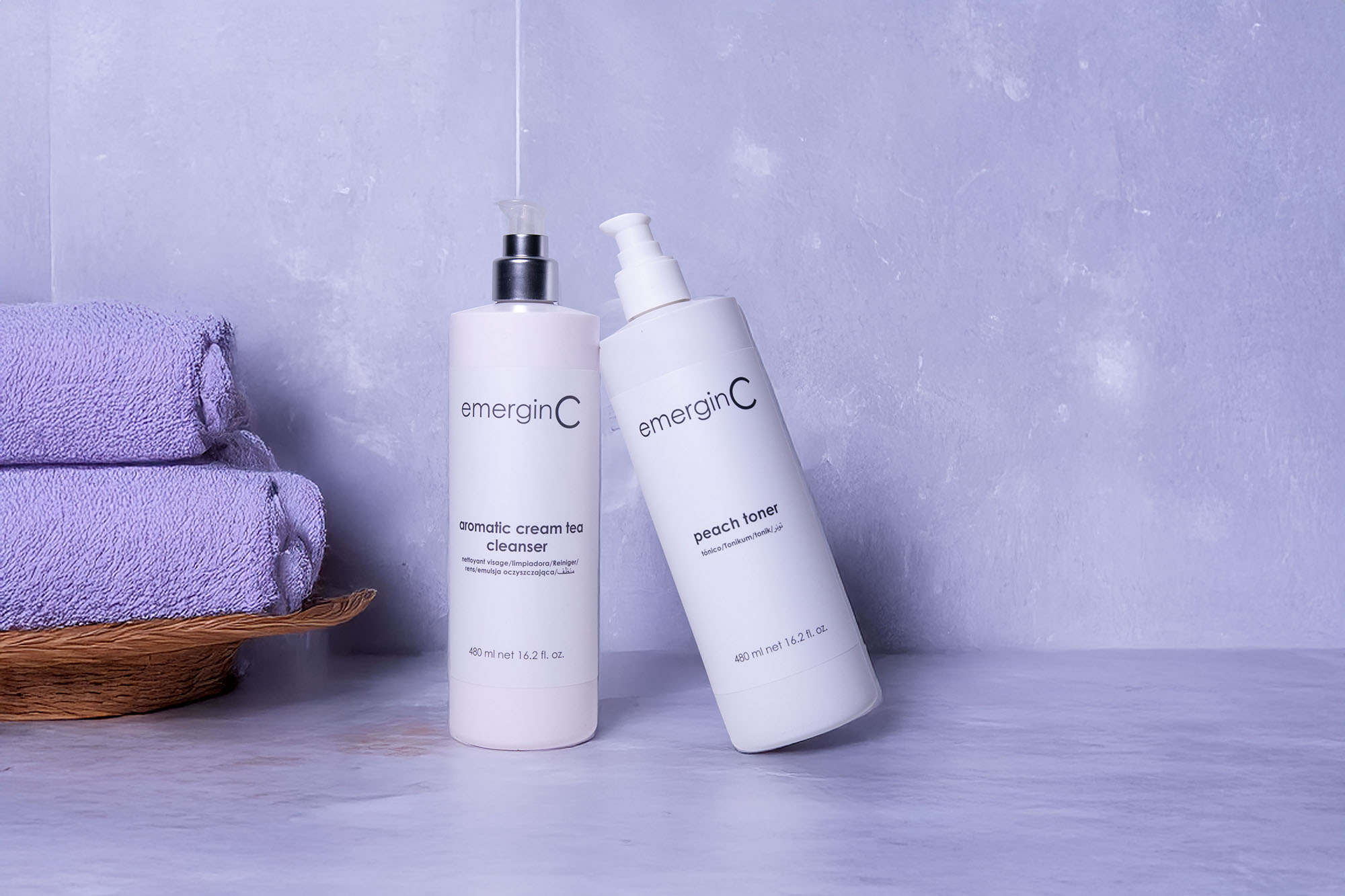 Two bottles of emerginC facial products, including an aromatic cream tea cleanser and peach toner, are placed on a light surface next to a stack of folded purple towels in a wicker basket against a light lavender background.