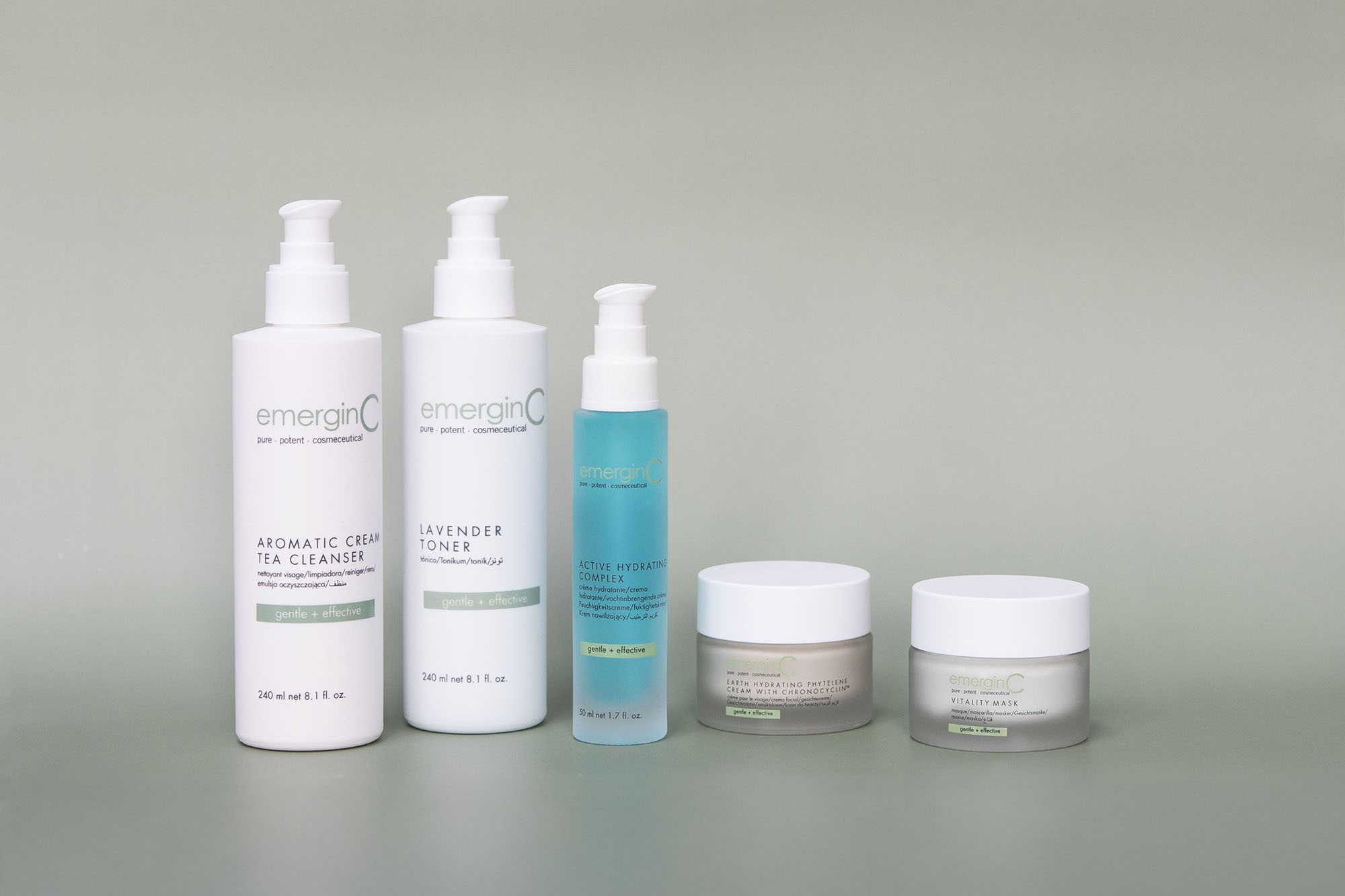 Five EmerginC facial products, including two pump bottles, a spray bottle, and two jars, are arranged side by side on a smooth, neutral-colored surface with a light green gradient background.