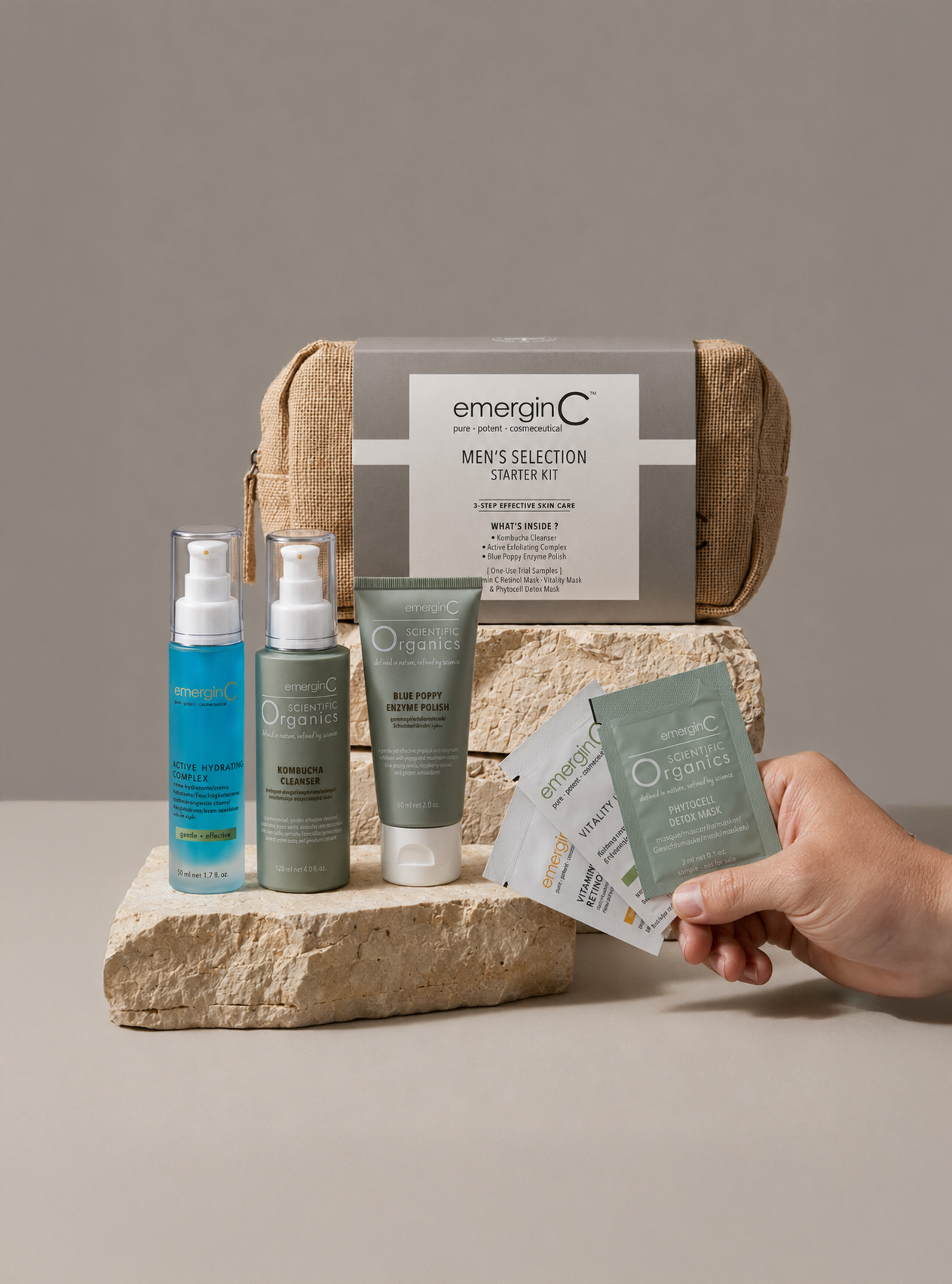 Professional luxury skin care products from the emerginC Men's Selection Starter Kit are displayed with a brown pouch, three bottles, and sample packets arranged on beige stone blocks against a neutral background.
