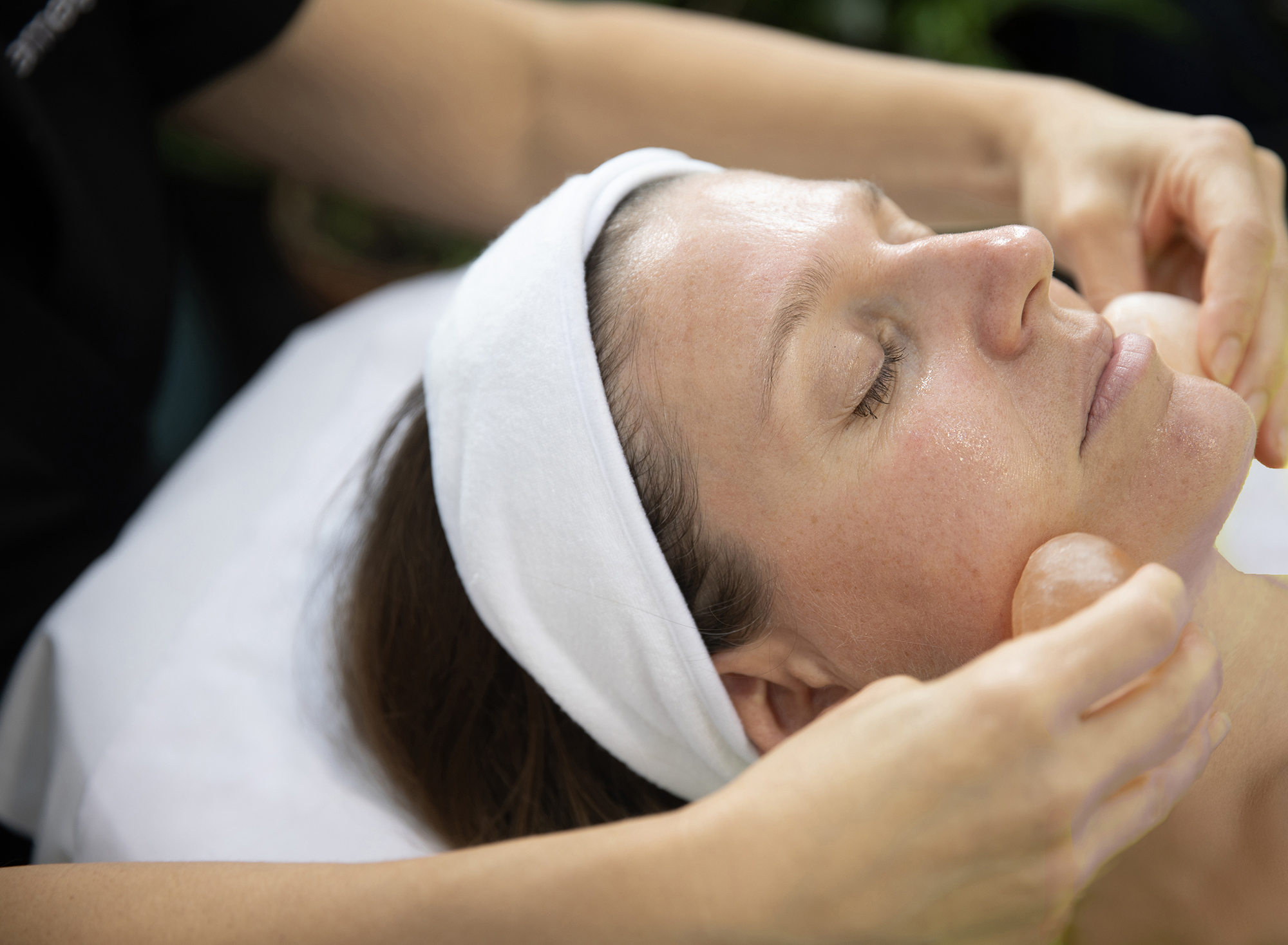 A person with closed eyes and a white headband receives a relaxing facial massage from another person's hands, suggesting a spa or self-care setting enhanced by emerginC facial products.