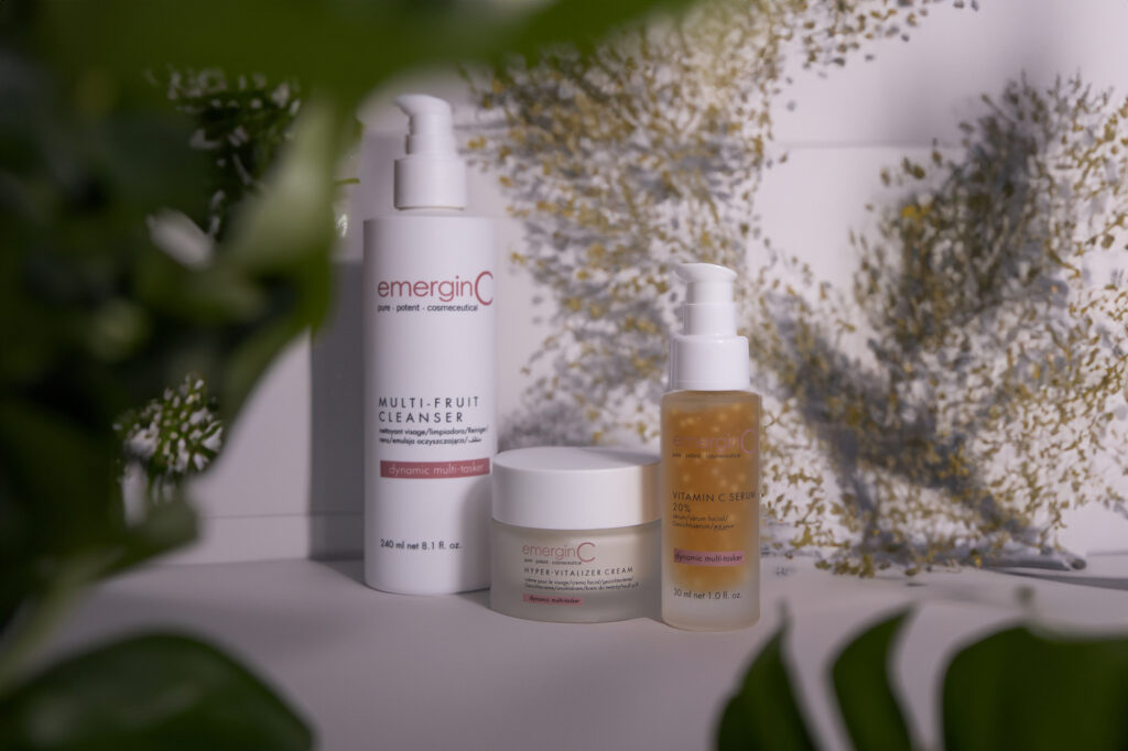 Three emerginC facial products—a cleanser, cream, and serum—are displayed on a white surface, surrounded by green leaves and delicate sprigs of tiny white flowers in the background.