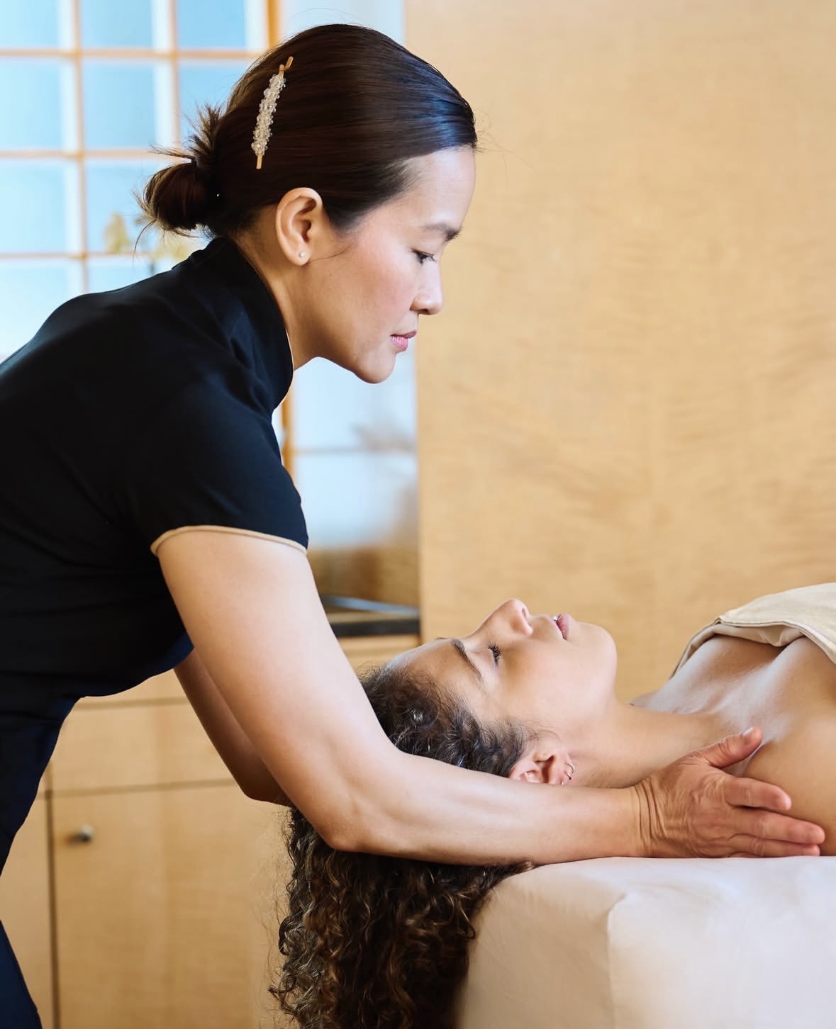 A massage therapist leans over a client lying on a table, supporting the client’s neck with her hands in a bright, serene spa setting.