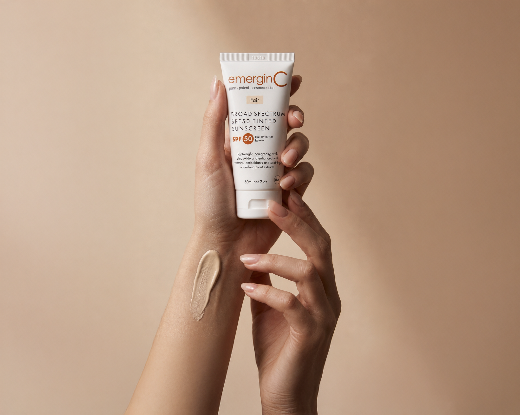 A hand holds a tube of emerginC SPF 50 sunscreen, while the other arm shows a streak of this professional luxury skin care applied to the skin, set against a beige background.