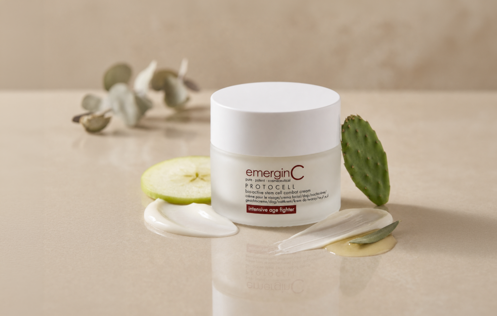 A jar of emerginC Protocell cream sits on a beige surface, surrounded by apple slices, cactus, green leaves, and cream smears, evoking a scene inspired by a skin care ingredients glossary and highlighting the natural elements within the formula.