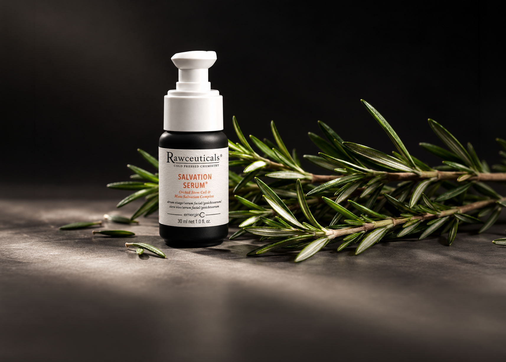 A bottle of Rawceuticals Salvation Serum, a professional luxury skin care product, stands beside sprigs of fresh rosemary on a dark surface, with dramatic lighting highlighting the serum.