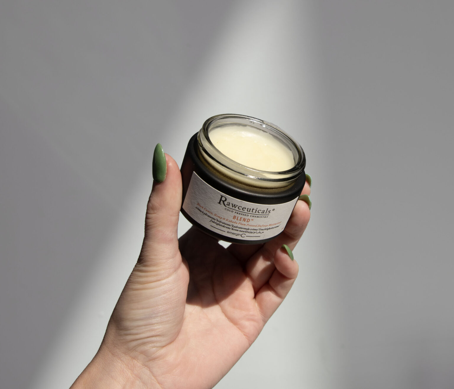 A hand with green-painted nails holds an open glass jar of Rawceuticals Blend face moisturizer.