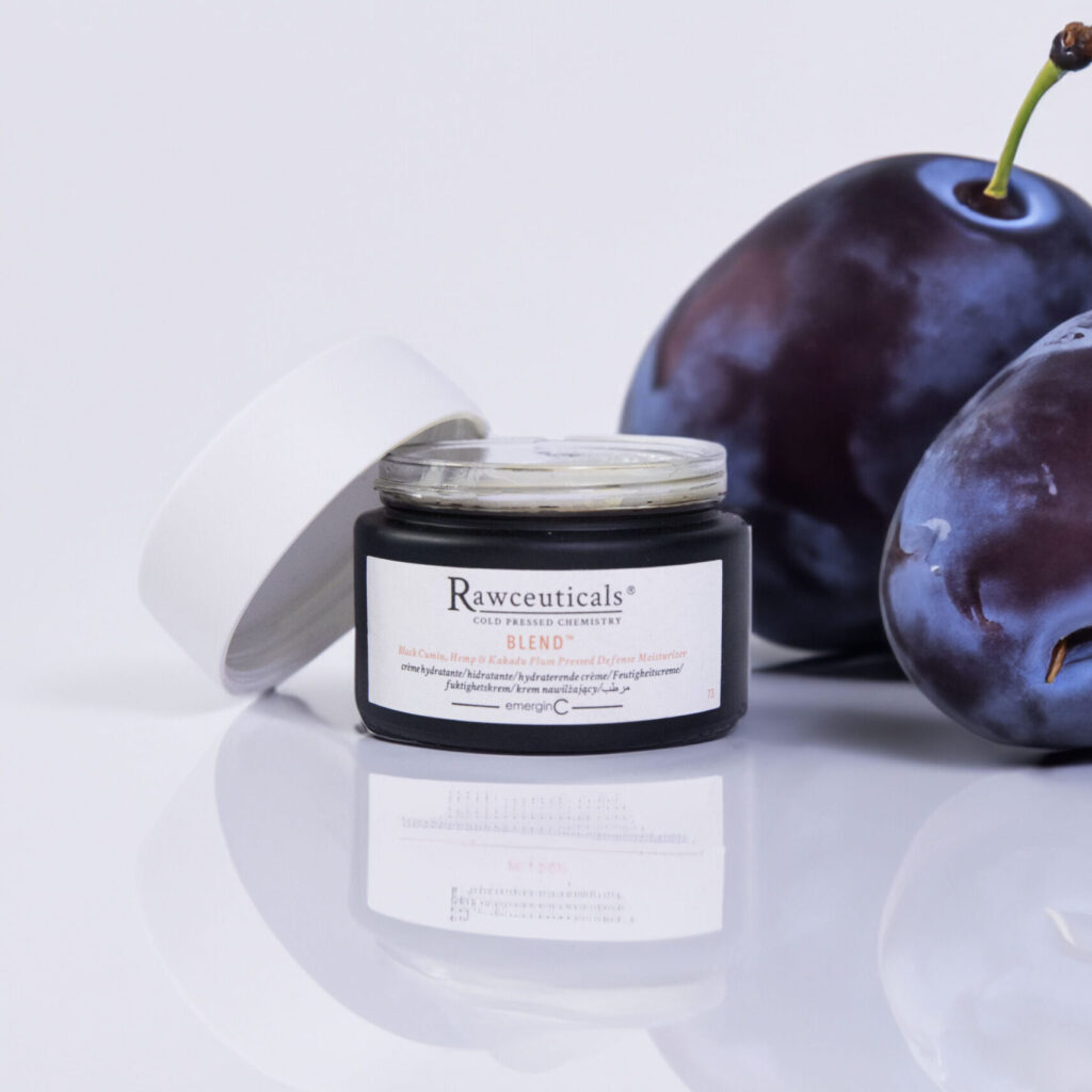 A jar of rawceuticals' BLEND™ skincare product elegantly presented beside two fresh, ripe plums on a clean, white surface, highlighting the theme of natural ingredients.