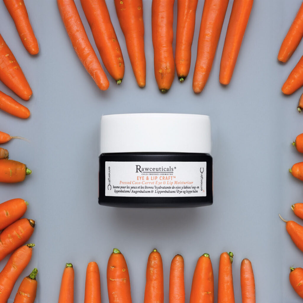 A jar of EYE & LIP CRAFT™ surrounded by a circle of fresh carrots on a blue background, suggesting the product contains carrot extracts or vitamins beneficial for the skin.