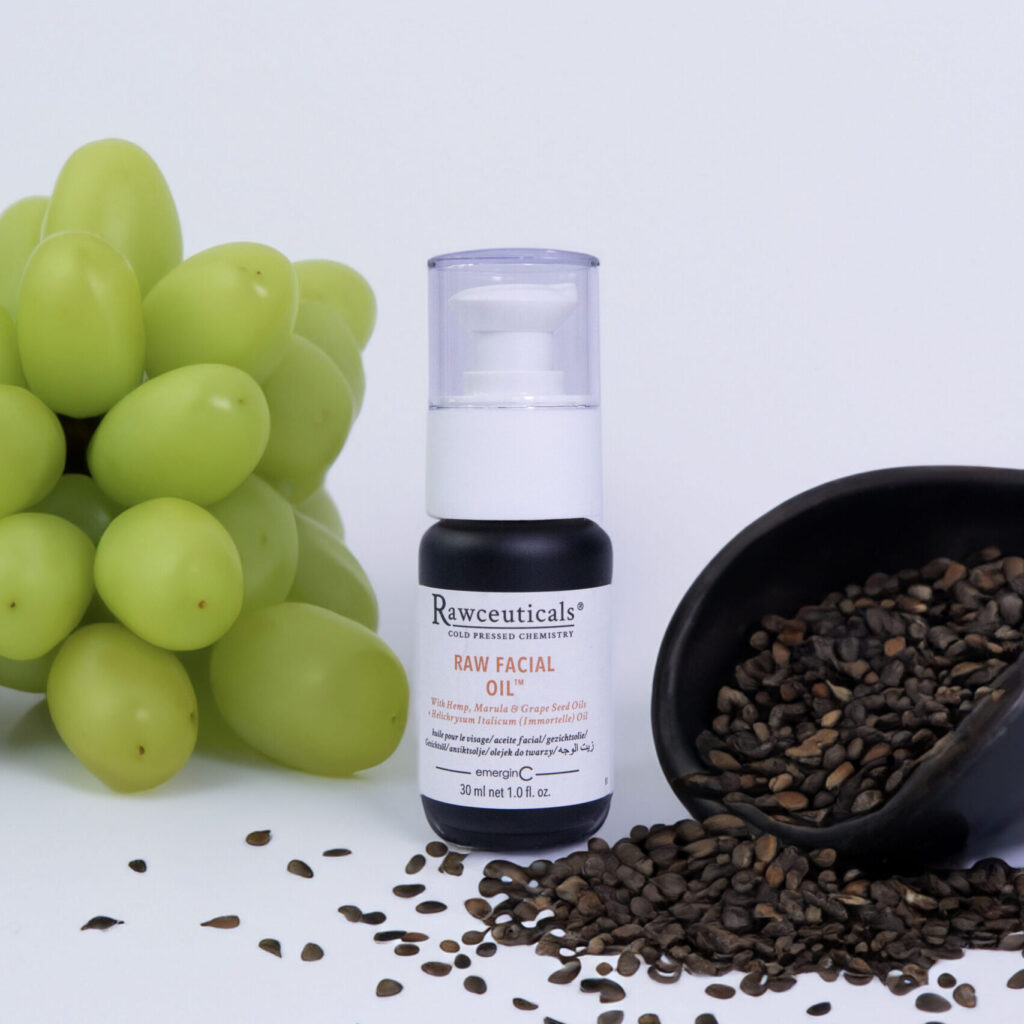 A bottle of RAW FACIAL OIL™ next to a bunch of green grapes with a scoop of scattered flaxseeds on a white background.